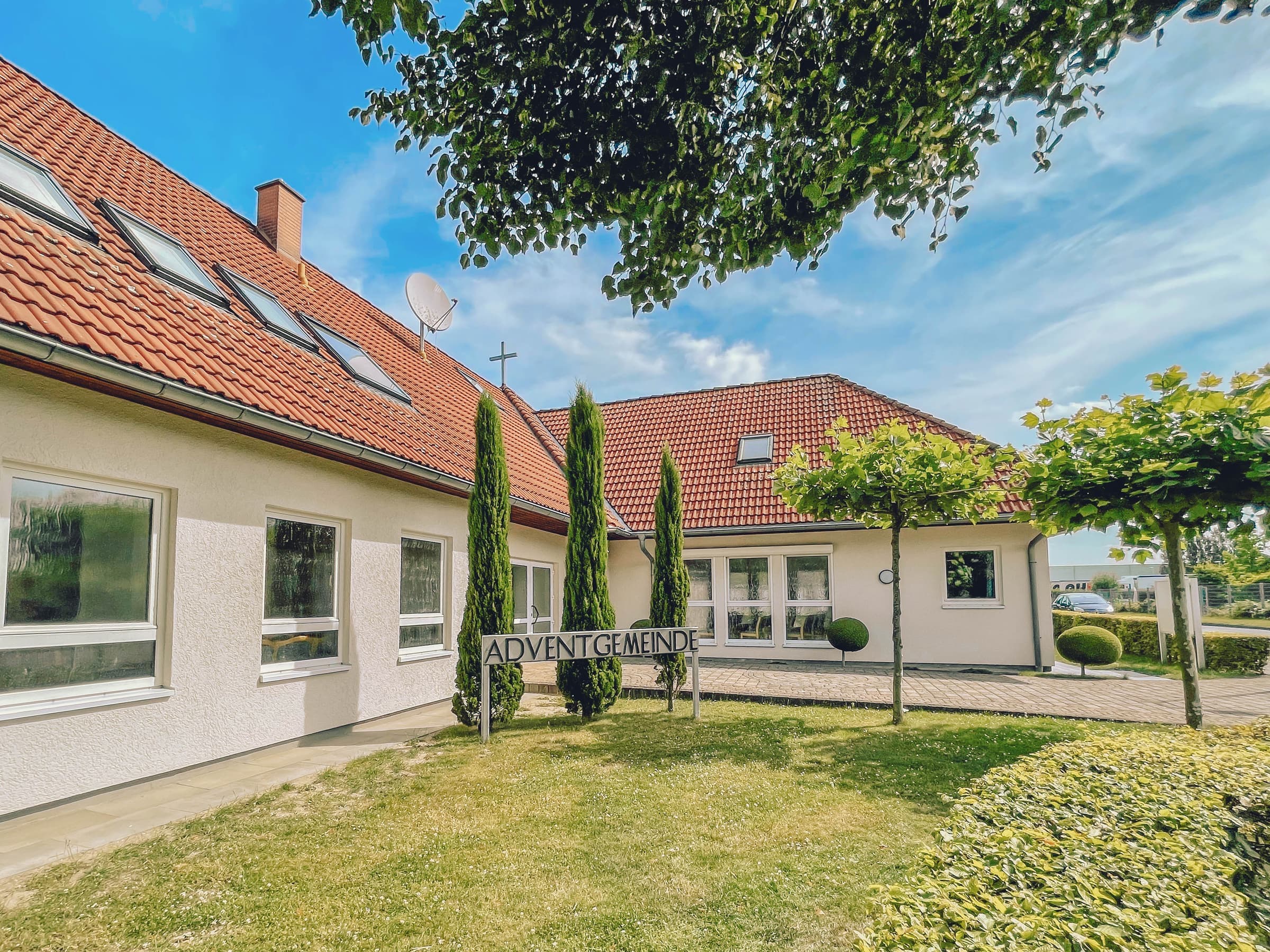 Das Gemeindehaus mit rotem Ziegeldach, davor im Rasen das Schild Adventgemeinde zwischen schlanken Zypressen